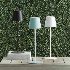 three lamps sitting on top of a wooden table next to a green plant covered wall