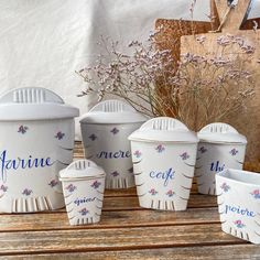 white ceramic containers with flowers on them sitting on a wooden table next to a potted plant