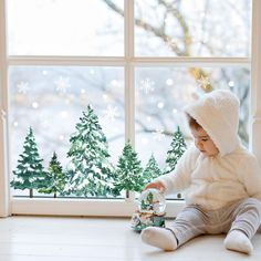 a baby sitting on the floor next to a window with snowflakes and trees