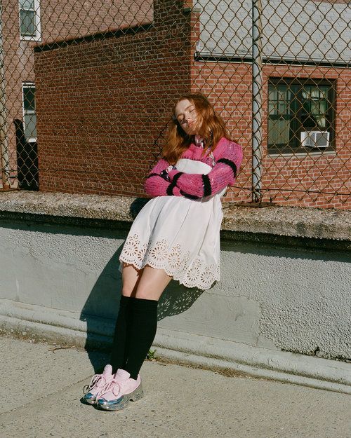 a woman in pink sweater and white skirt leaning against a wall