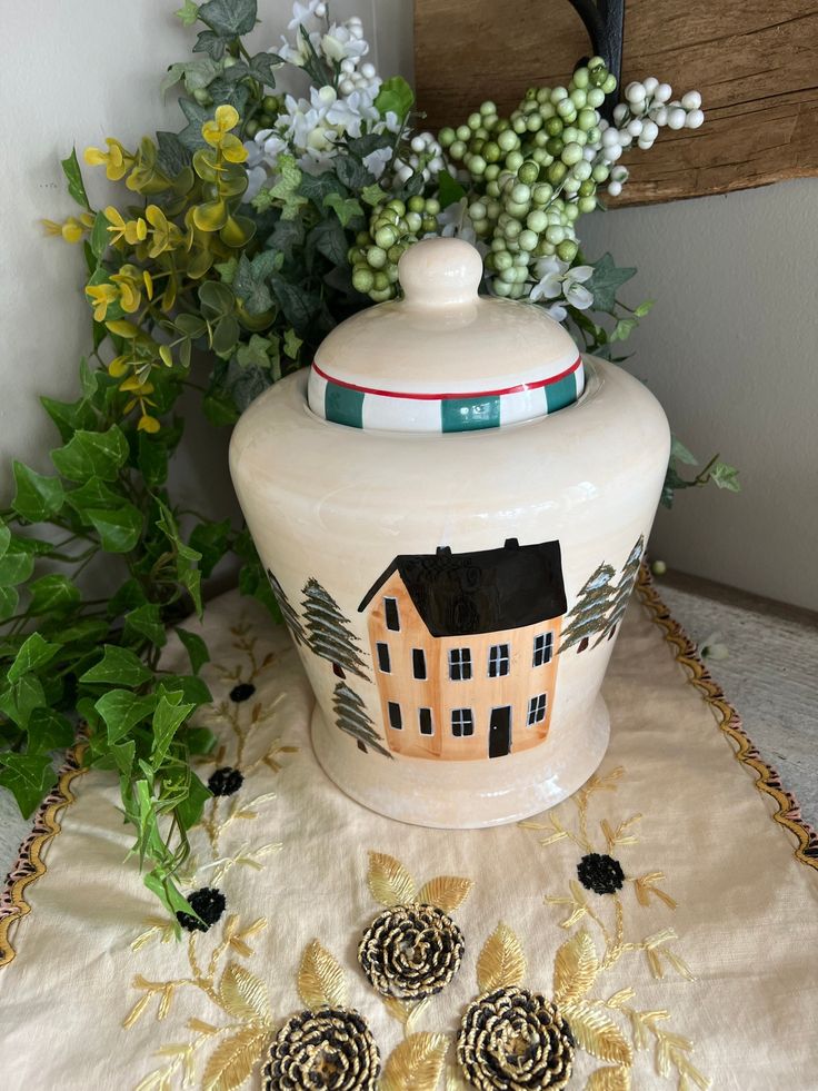 a white vase with a house painted on it next to some flowers and greenery