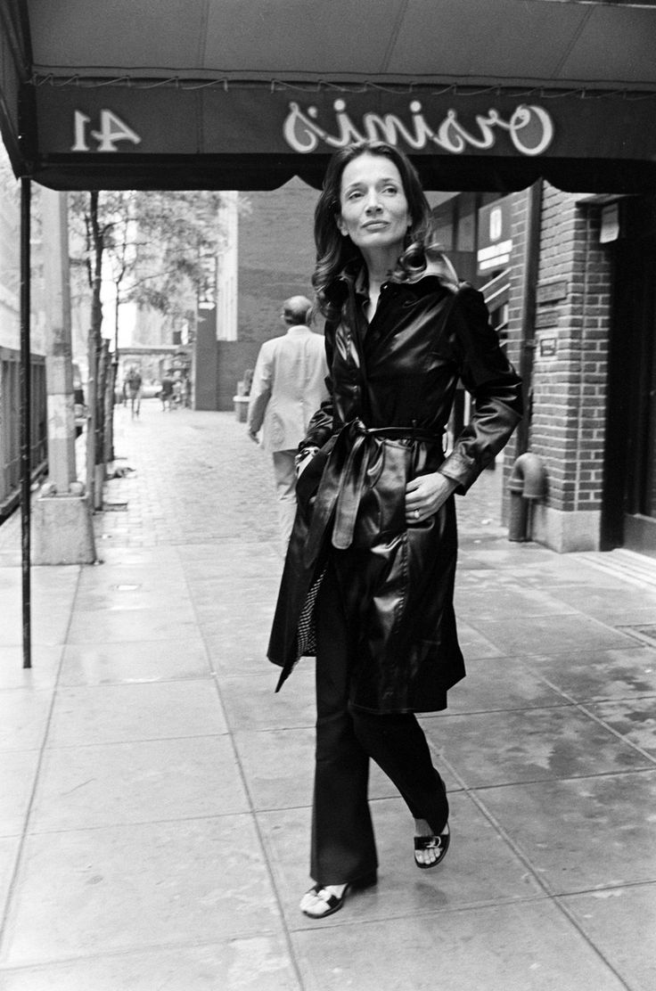 black and white photograph of woman in trench coat walking on sidewalk near business name sign