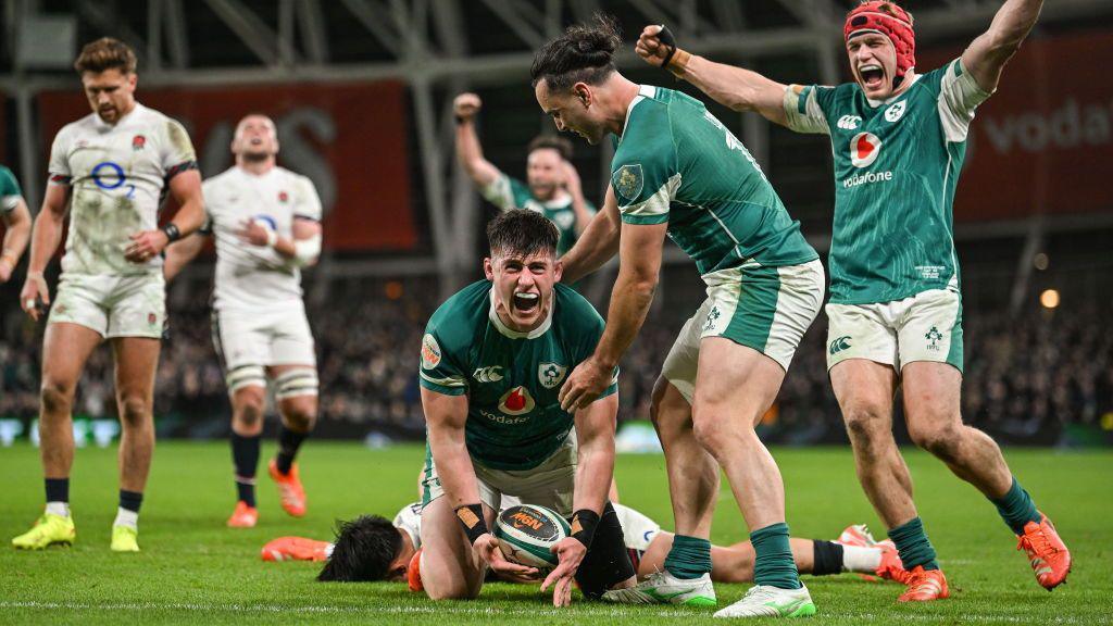 Three men dressed in green shirts cheering on a rugby pitch