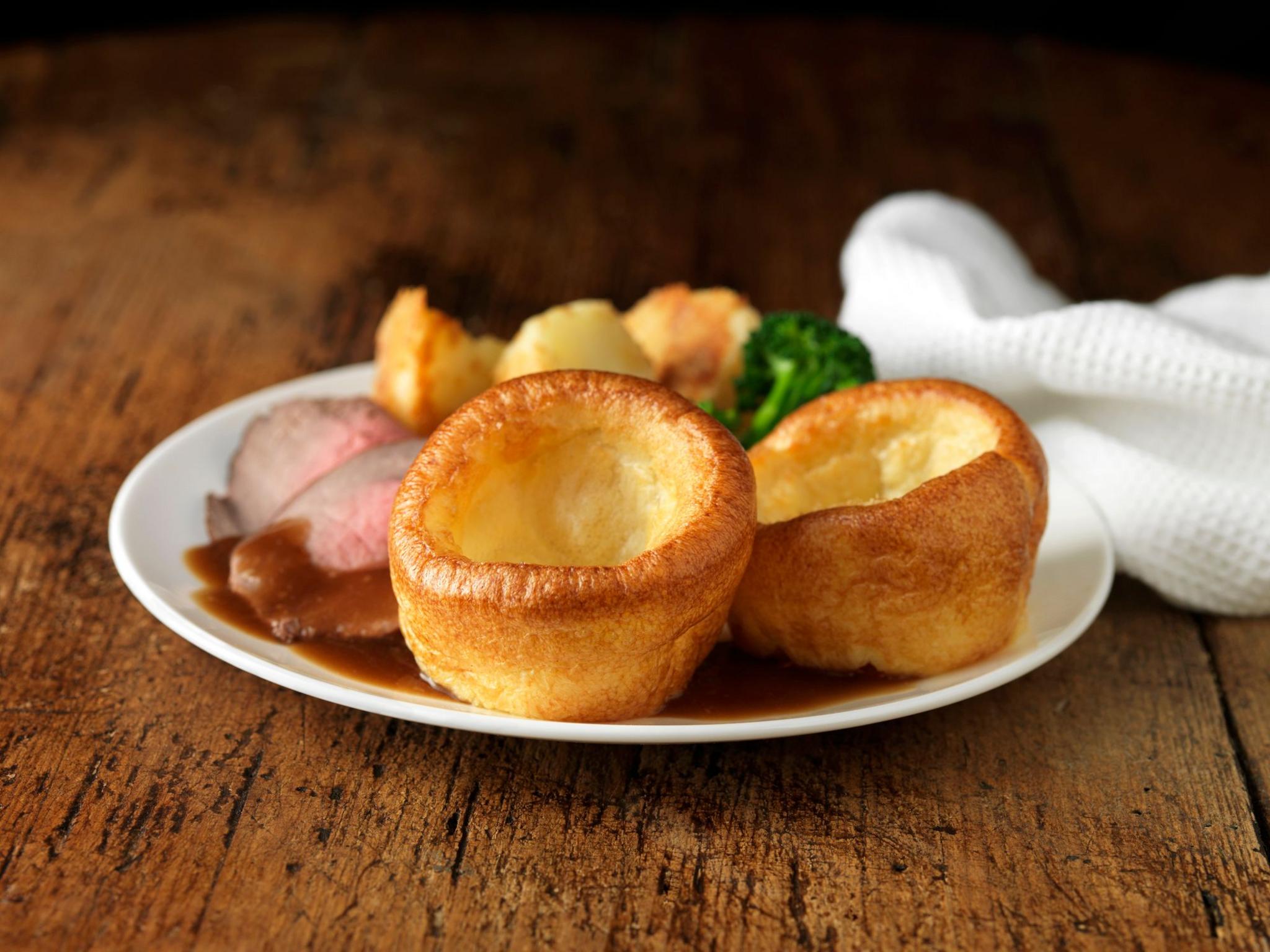 two yorkshire puddings sat on a plain plate with gravy, roast meat, roast potatoes and broccoli - the plate rests on a wooden table with a white cloth next to it