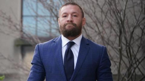 Conor McGregor walking into court. He is wearing a blue suit jacket with a white shirt and a navy tie.