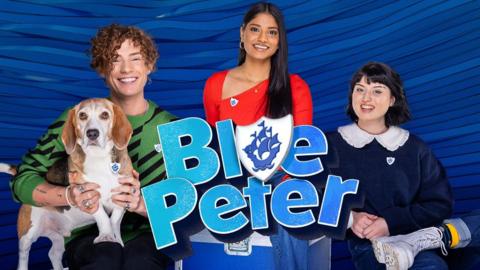 The Blue Peter presenters and Henry the dog, from left to right there is Henry, a beagle basset hound cross dog, Joel, a young white man, Shini a young Tamil woman, and Abby a young white woman.