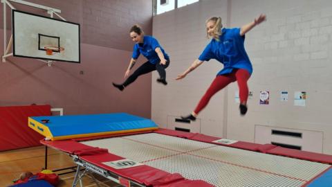 Two women in blue t-shirts mid air following a jump on a wide trampoline with red and blue trim. 