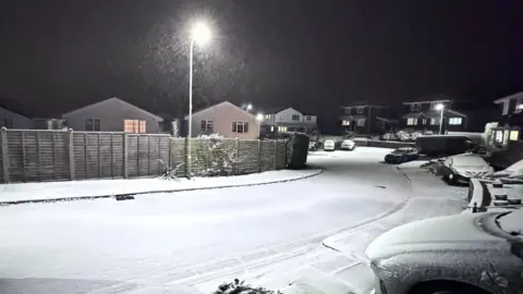 A snowy street in Wales. There are cars lining the suburban road and houses can be seen in the distance. It is night time. Snow is falling and coats the ground.