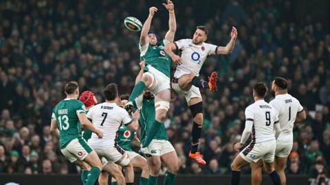 Two rugby players wearing a green Ireland and a white England shirt jump for the ball with other players around them 