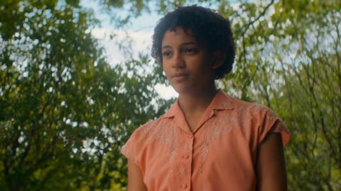 A young black girl with short black curly hair is wearing a salmon-coloured shirt and is standing in a forest