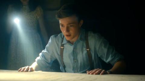 A young white boy is looking at a table lit by a girl holding a torch. The boy has short brown hair and is wearing a blue shirt with brown suspenders