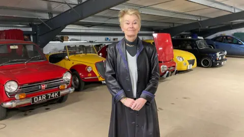 A woman with short blonde hair stands in front of a row of classic cars of a variety of colours. She is wearing a long black leather coat.