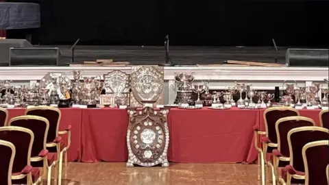 A long red table with multiple brown shields with silver-plated inscriptions. there is also a host of silver trophies on the table, which is in front of a stage. Gold and red chairs have been set out in rows nearby.