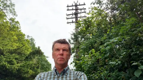 Martin Evans, with short brown hair and wearing a blue and brown checked shirt is standing in front of a telegraph pole, staring ahead in the distance. There are trees either side of him