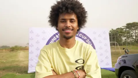 Karim Adeyemi smiles into camera as he stands with arms folded in yellow shirt