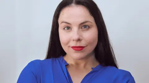 A woman with long dark hair is smiling at the camera. She is stood in front of a white background and is wearing red lipstick and a blue blouse.