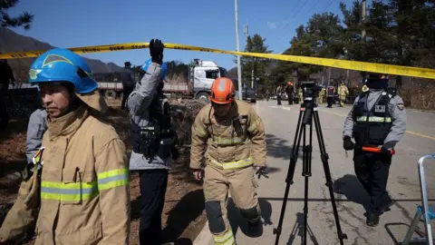 Firefighters and security personnel crossing under a yellow police tape cordoning off the bomb site