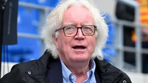 Winfried Schafer, wearing a black jacket over a blue and white striped shirt, is seen in front of a stand of blue seats