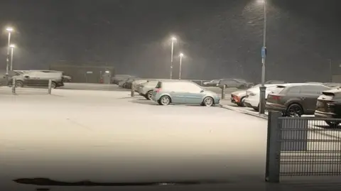 Heavy snow is falling in a car park where a number of cars are pictured.