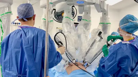 A photo from the operating theatre. It shows two surgeons standing with their back to the camera, wearing blue surgical gowns. They each have a hand on one of the robot's surgical arms. The four robot arms are inserted into the abdomen of a patient who lies on an operating table. We can only see the patient's abdomen.