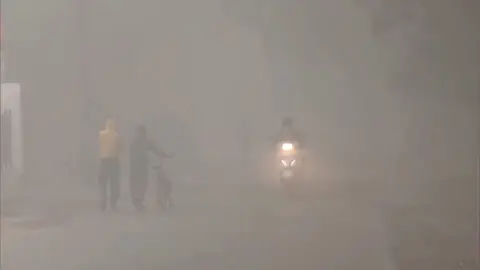 A couple is seen walking with a bicycle while a man on a scooter passes them in dense fog