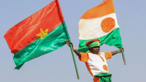 Men hold a boy up in the air. He is waving the flag of Burkina Faso and the flag of Niger.