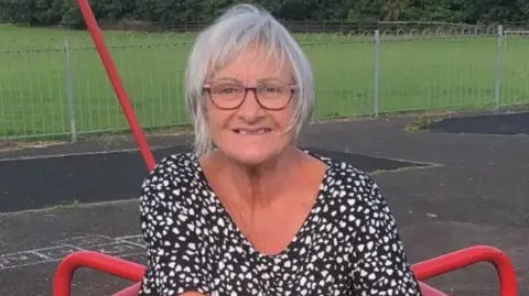 Elizabeth Kennedy sitting on a piece of red playground equipment. She is looking directly at the camera. She has grey hair, which is partially obscuring her face. She is wearing a black top with a small white heart pattern covering it.