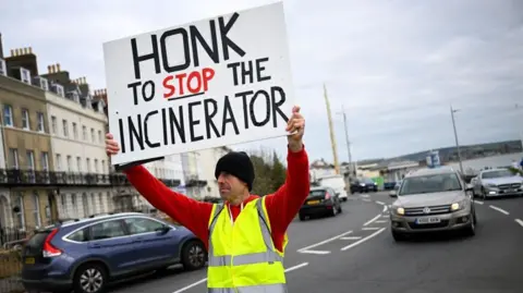 A man in a hi-vis jacket holds a sign that reads: Honk to stop the incinerator. The word stop is underlined. He is standing in the middle of the road facing the camera, with a car behind him, and a row of houses to his right.