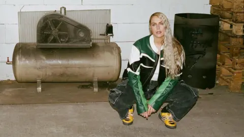 Sally is crouched down beside a generator. She is wearing a green, white and black leather jacket. She has long blonde wavy hair. She is also wearing bright yellow shoes. 