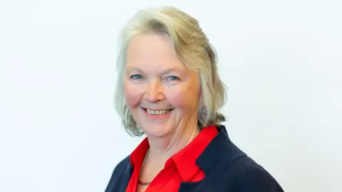 Councillor Jane Meagher looking directly at the camera. She has white-blonde hair. She is wearing a red shirt under a black jacket.