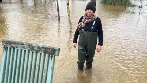 A woman in waders is walking through floodwater which reaches up to her mid-shin. There is a turquoise piece of garden furniture in the foreground. 