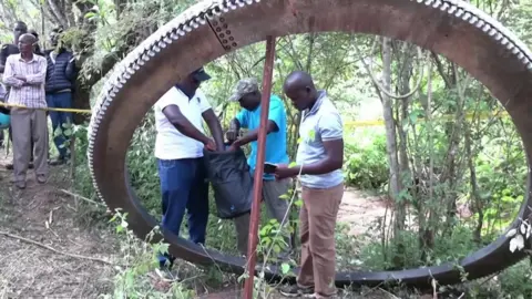 Three men in T-shirts are standing inside a huge metal ring as they investigate what happened. Onlookers can be seen at the back of the picture.