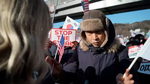 A pro-Yoon supporter holding a US and South Korean flag in each hand speaks to Jean Mackenzie.