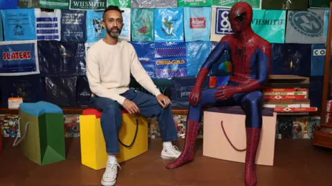 Hetain Patel is sitting on a stool that looks like a yellow shopping bag. He has short dark hair and dark and grey facial hair. Sitting next to him on a similar stool is a life-size model of Spider-Man in a blue and red suit. In the background is a wall made up of branded plastic bags.