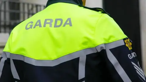 An officer in a Garda uniform. Shoulders and upper back are visible. They are wearing a yellow high vis jacket wiht silver stripes and black. There is a Garda logo on the right arm. The word 'Garda' is written on the back.