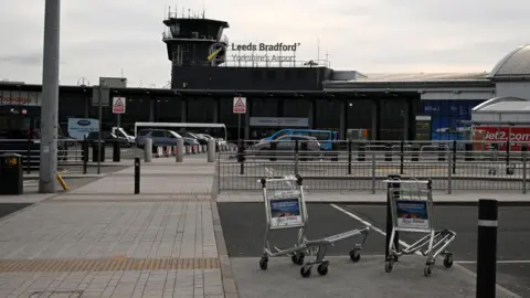 The outside of Leeds Bradford Airport. The airport appears to be deserted. There are two luggage trolleys abandoned by the side of a pavement leading up to the site entrance. 