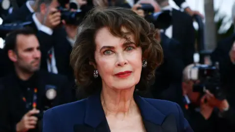 Dayle Haddon looks to camera. She is standing on a red carpet, with photographers behind her 