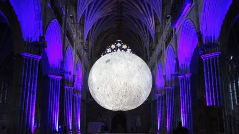 A large spherical sculpture of the moon hangs in the arches of a gothic cathedral at night, with the columns illuminated by purple light.