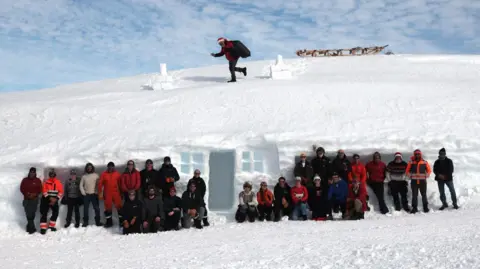 Group of around 30 people in front of a snow house with someone dressed as Santa on the roof.