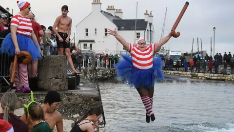 A woman dressed in a 'Where's Wally' costume holding an inflatable walking stick, jumps into the water as people stand and watch from the habourside. January 1, 2025 