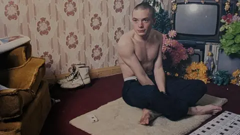 A photograph from the exhibition shows a young man, with cropped hair and no shirt sitting on the floor in a 90s living room