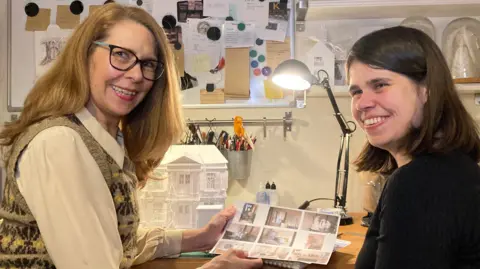 Jacqui and Lauren Saunders-Love, sitting in a studio, smiling at the camera. Jacqui, to the left is holing images of artwork, wearing a cream top and tank-top, Lauren has on a black top. There are art times, all around them, a noticeboard and black lamp. 