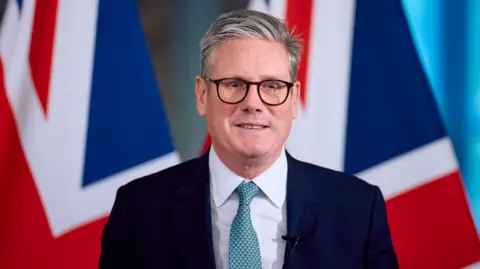  Britain's Prime Minister Keir Starmer speaks at a press conference, during his visit to the European Commission headquarters on October 2, 2024 in Brussels, Belgium. Behind Sir Keir are two flags of the UK in the distance. He is wearing a navy suit, black spectacles and a blue tie.