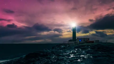 The lighthouse is centre of shot, burning bright against the purple and orange aurora. The light is reflected in puddles on the shore in the foreground.