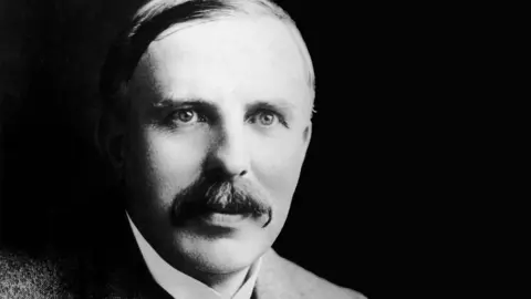 Sir Ernest Rutherford, with a large moustache, side-parted hair and a stiff white collar, stares past the camera