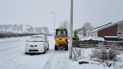 A silver car covered with snow is parked at the side of the road in Balerno. A grit spreading tractor is next to it on the pavement. It is yellow with a red and yellow banner and the word "spreading" in red text. A driver is visible in the cabin wearing an orange hi-vis vest. The ground, including the road, is almost completely covered in snow. The sky is grey and the trees in the background and foreground are bare.