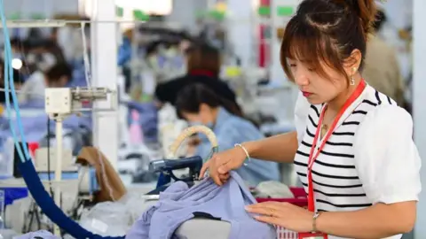 A woman in a white top, wearing a red lanyard, works on a sewing machine which has a blue top on. Blue wires trail from the sewing machine