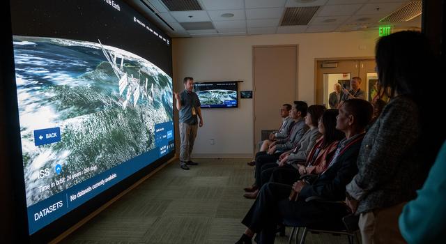 A man standing in front of an audience points to a large screen showing a simulated view of the ISS orbiting Earth.