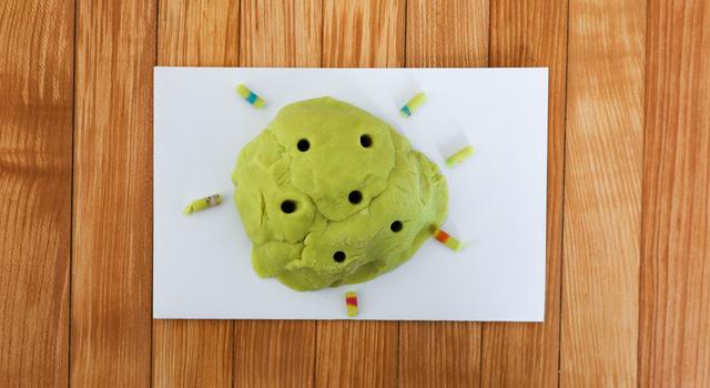 A green play dough rock is shown with multiple cores taken out of it and sitting next to it on a table.