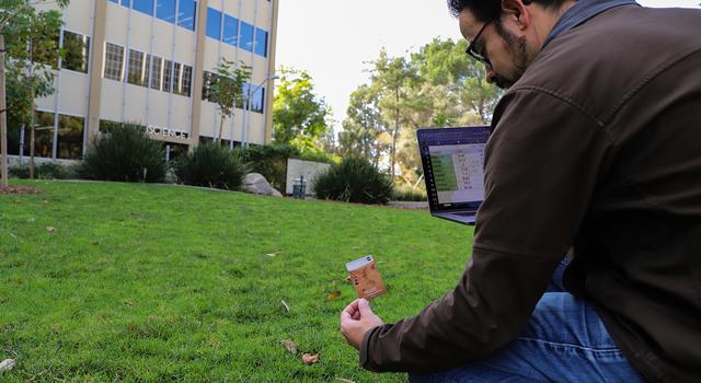 A man holds a computer with a spreadsheet of scale distances to the planets and places a Mars marker in the grass.